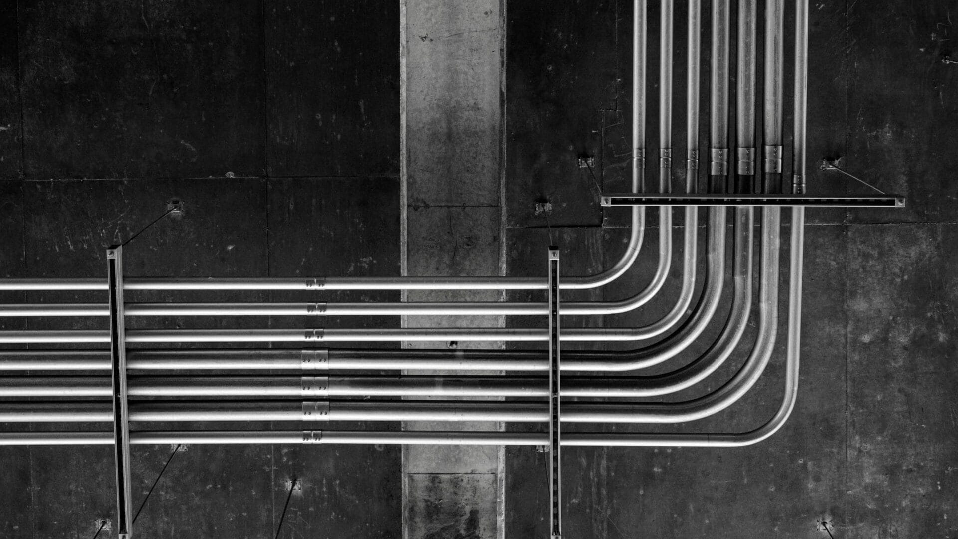 A black-and-white photograph showing several metal pipes running parallel and then curving upward, mounted on a dark concrete wall.