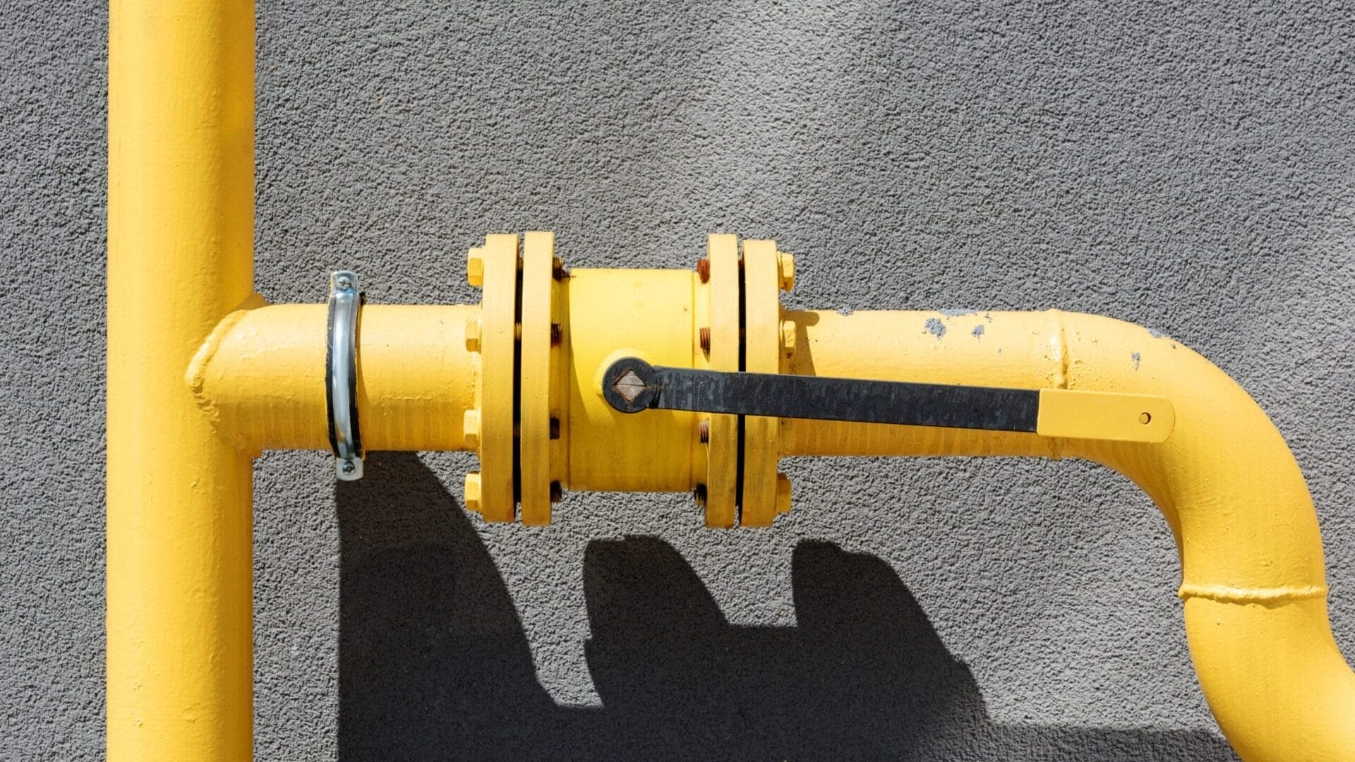 Close-up of a yellow industrial pipe with a black metal lever handle against a textured gray wall, certified by RPZ Valve Testers.