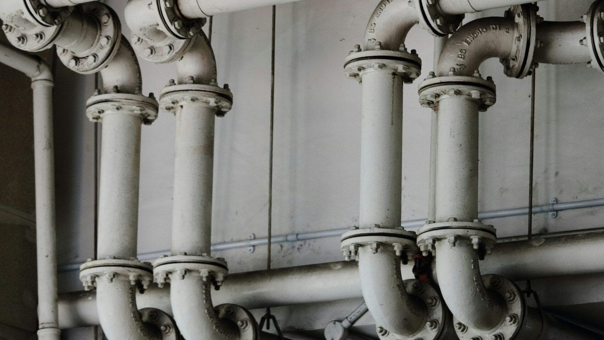 A network of white industrial pipes and fittings runs horizontally and vertically against a white wall.